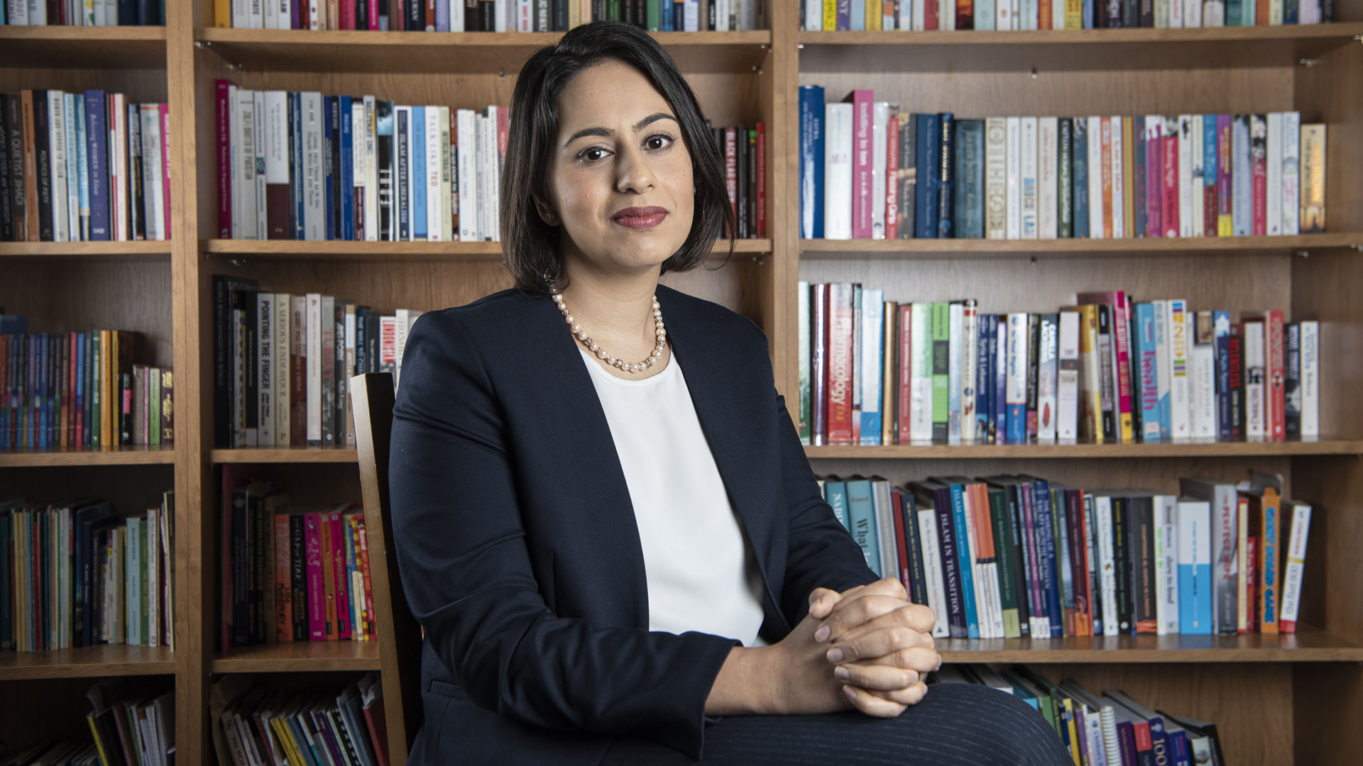 Sara Khan wearing black suit, smiling and sat in front of bookshelves full of books