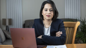 Sara Khan with arms folded, sat at a wooden desk with a rose gold laptop, wearing a black suit jacket